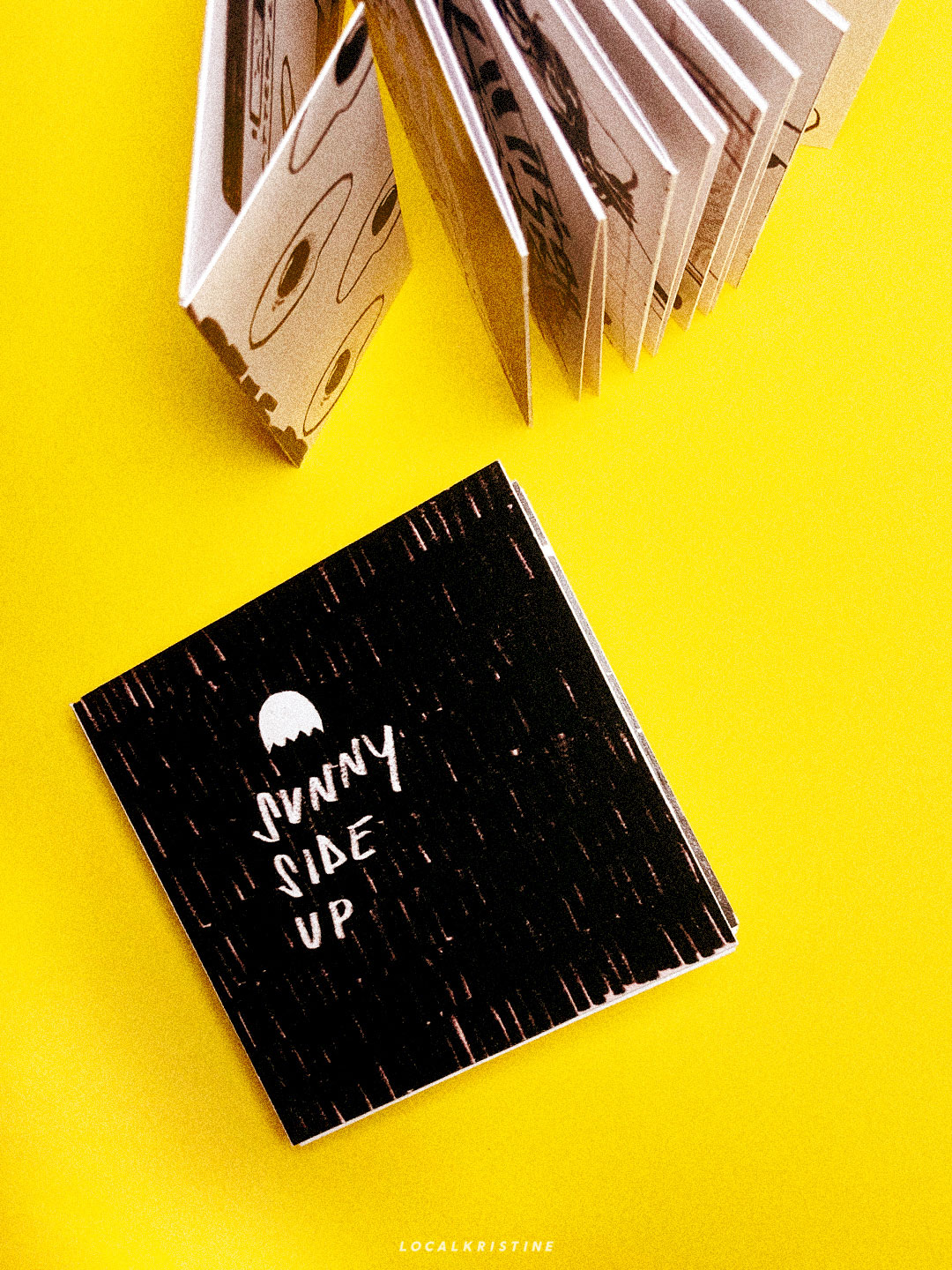 Overhead shot. Two zines. One lays flat and reads: Sunny Side Up. The other is standing with pages open. Book jacket has eggs on it.